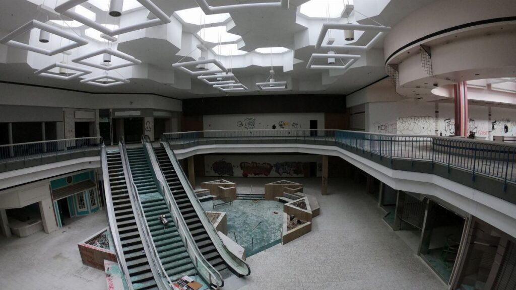 Desolate interior of an abandoned shopping mall with broken escalators and graffiti-covered walls.