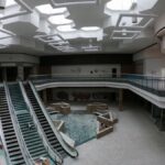 Desolate interior of an abandoned shopping mall with broken escalators and graffiti-covered walls.