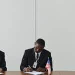 Executives signing international agreement with EU and US flags displayed on a wooden table.