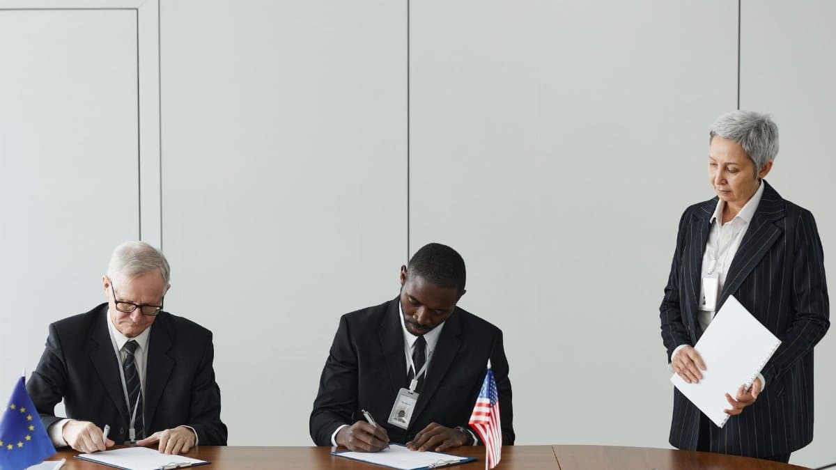 Executives signing international agreement with EU and US flags displayed on a wooden table.