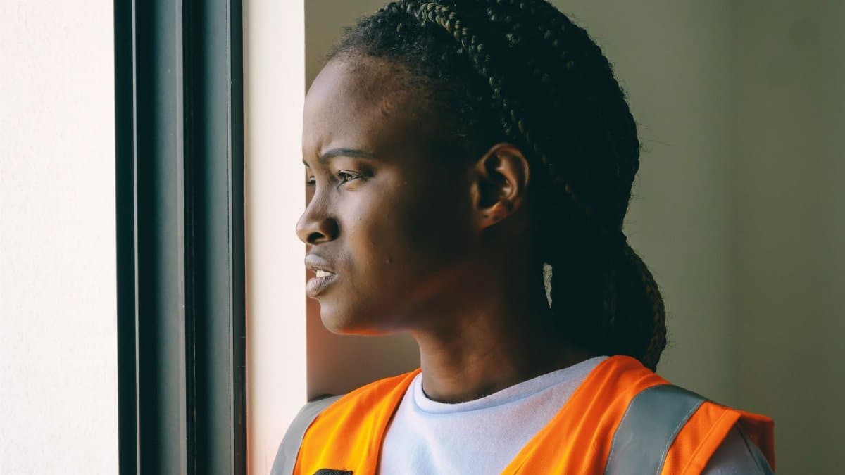 Profile of an adult African American woman wearing a reflective orange vest, looking out a window.