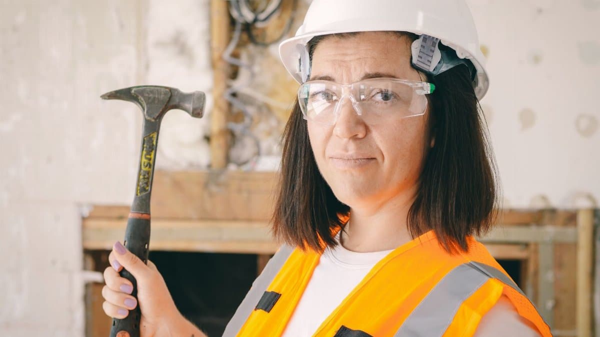 Confident female construction worker holding a hammer, equipped with safety gear including a hard hat and reflective vest.
