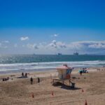 Panoramic view of a sunny day at Huntington Beach, featuring a lifeguard tower, people enjoying the beach, and ships on the horizon.