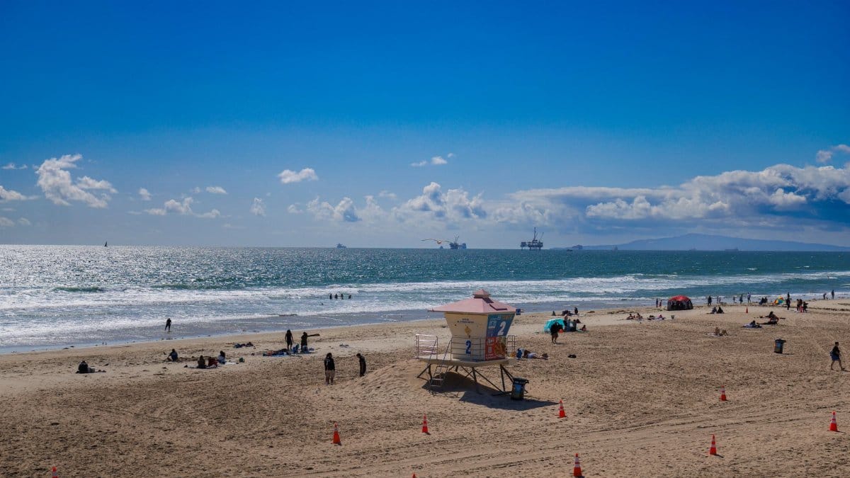 Panoramic view of a sunny day at Huntington Beach, featuring a lifeguard tower, people enjoying the beach, and ships on the horizon.