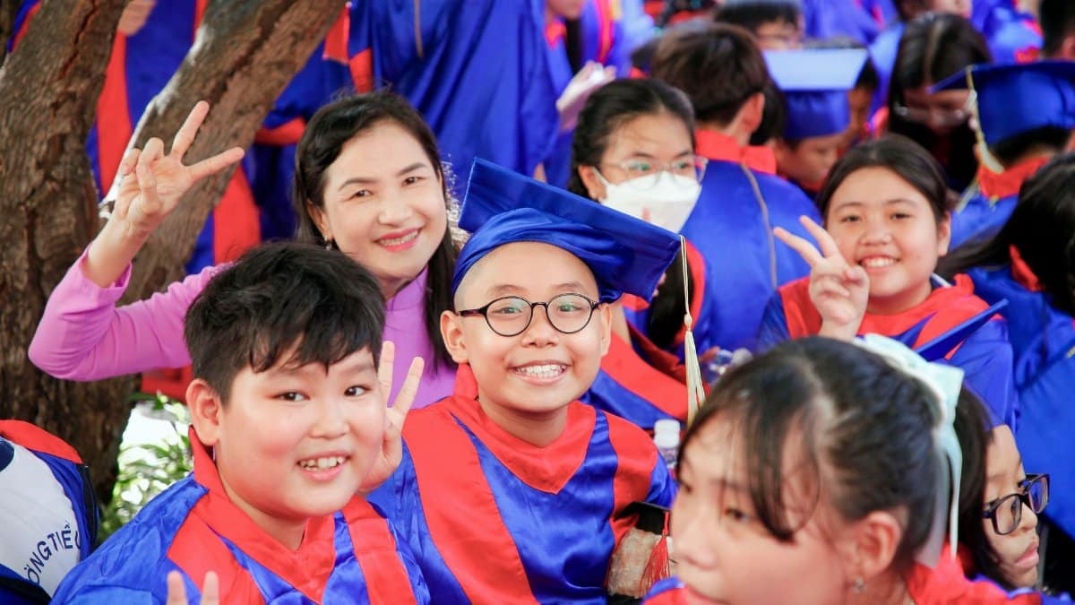 Group of children in colorful gowns celebrating graduation outdoors with smiles and peace signs.