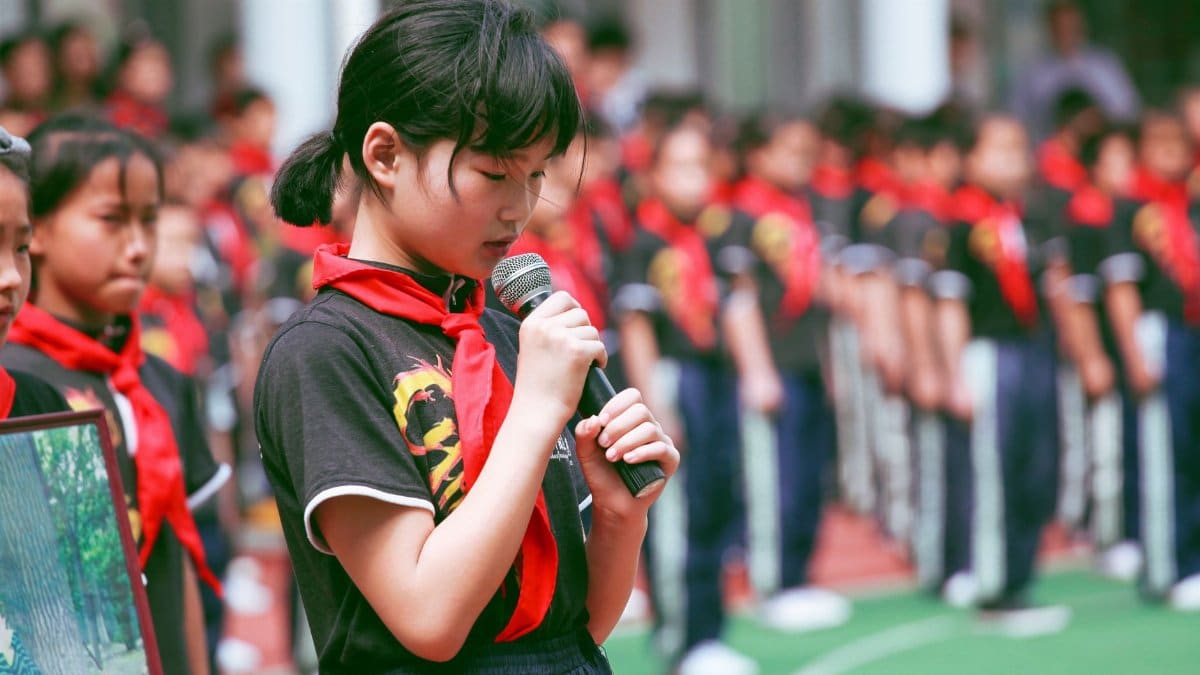 A young girl in uniform with a red scarf speaks into a microphone during an outdoor school event.