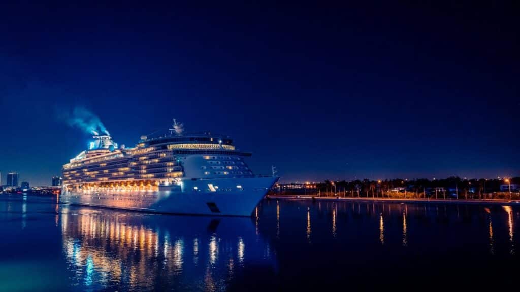 A brightly lit luxury cruise ship sails through Miami's waters at night, reflecting in the calm sea.
