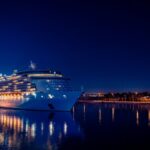 A brightly lit luxury cruise ship sails through Miami's waters at night, reflecting in the calm sea.