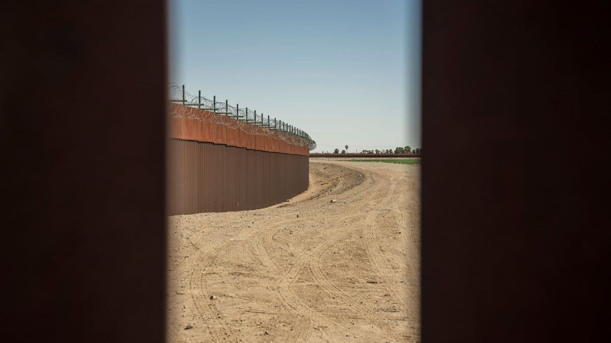 View of a barbed wire border wall structure alongside a dirt road under a clear blue sky.