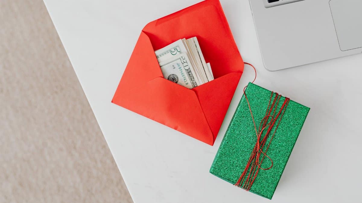 A green gift box and red envelope with cash on a desk beside a laptop, emphasizing holiday or financial themes.