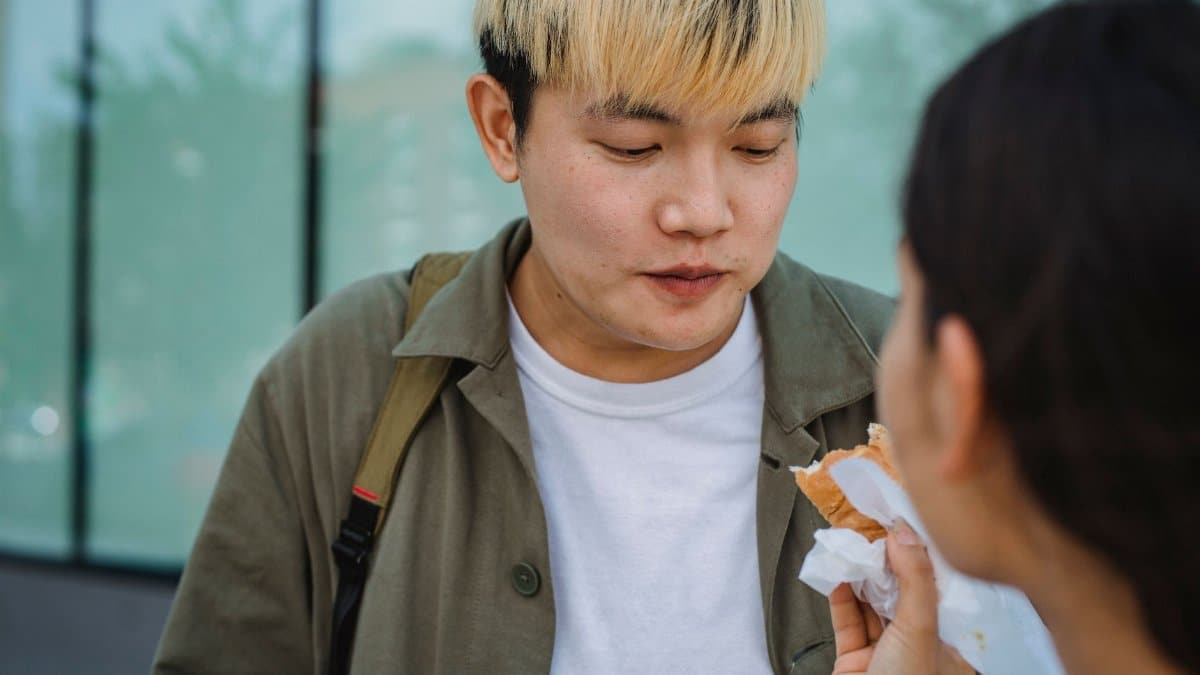 Crop female hand feeding young Asian boyfriend in casual summer wear with delicious hot dog while standing together on street