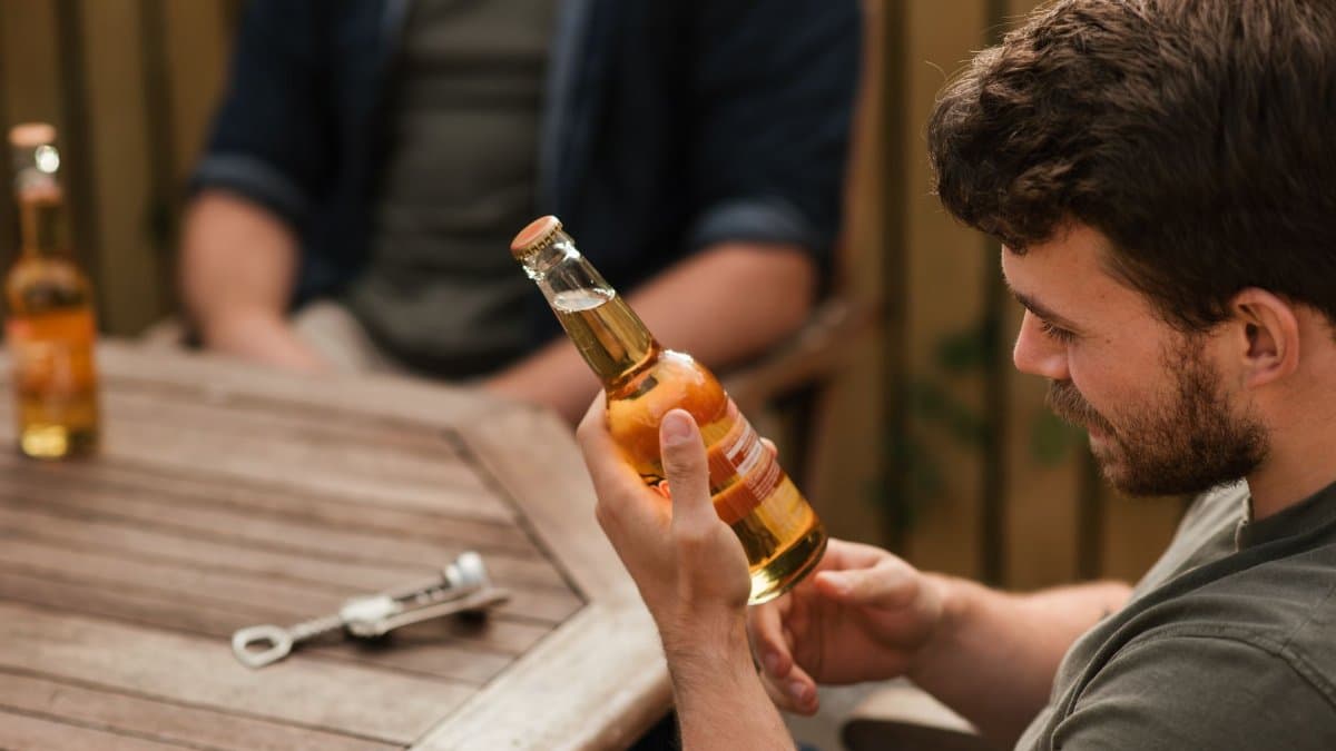 From above of crop bearded male reading label on glass of beer while sitting at table with friend in backyard