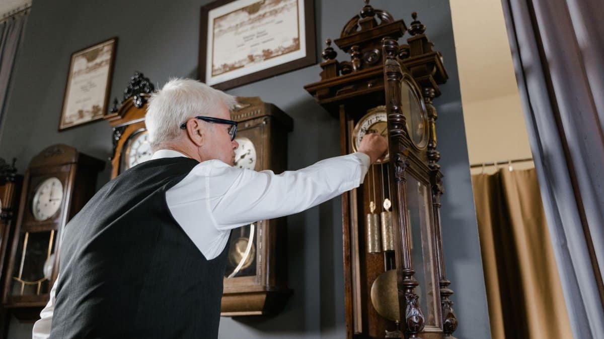 An elderly man adjusts an antique grandfather clock inside a vintage clock shop.
