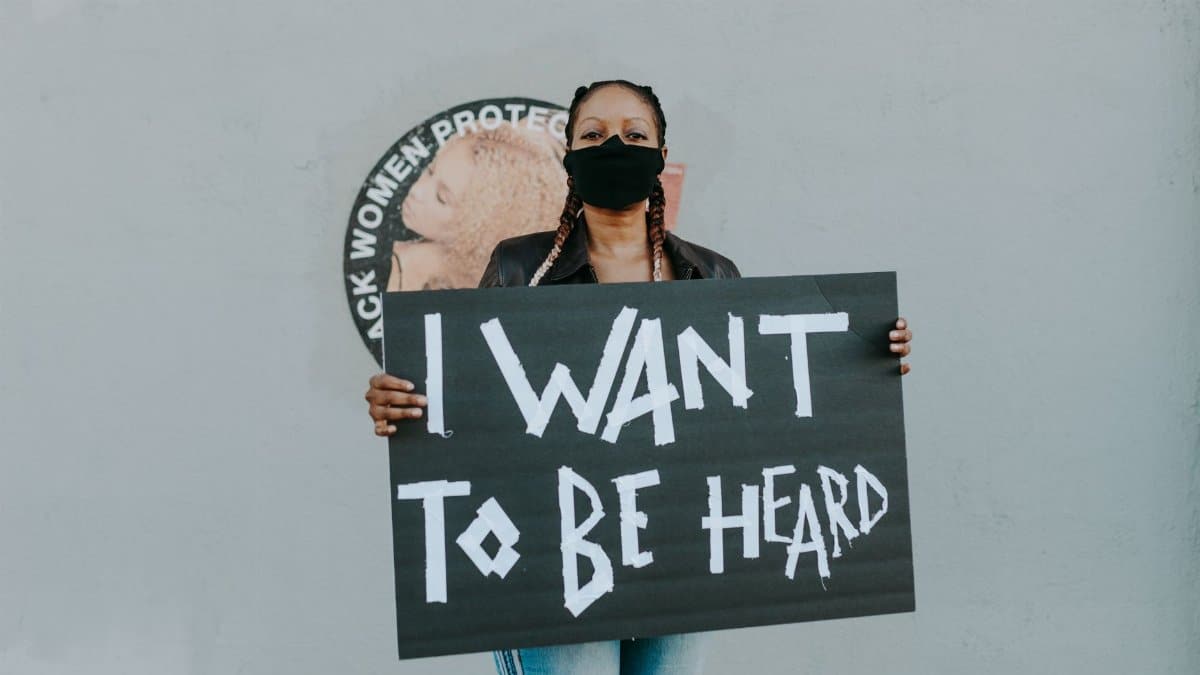 Masked protester holds a sign saying 'I Want to Be Heard' against a wall with activist imagery.