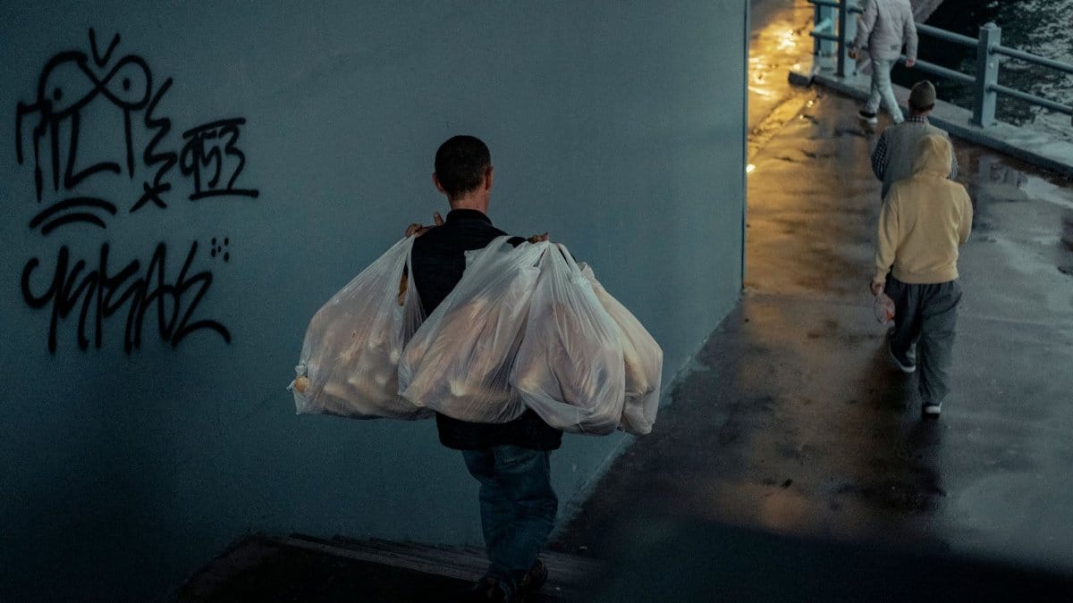 A person carrying plastic bags walks past graffiti in a dimly lit city street.
