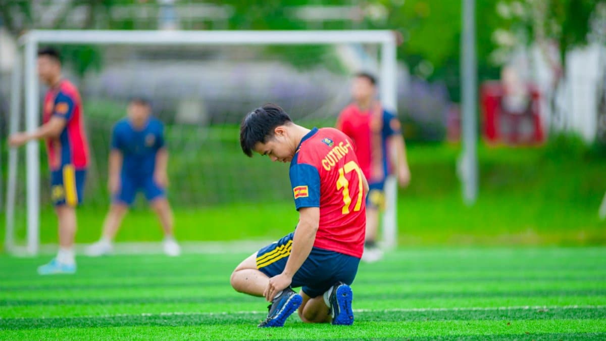 Young football player ties shoe on a sports field in Hanoi, Vietnam with teammates in the background.