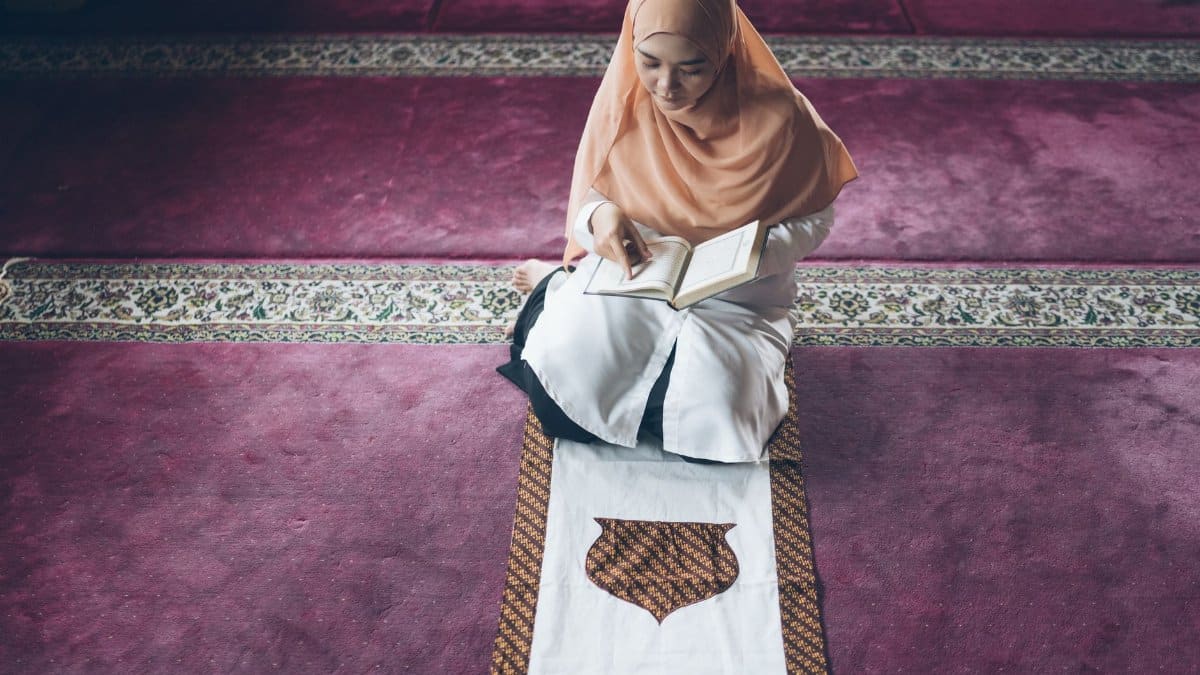A Muslim woman in a hijab reading the Quran on a prayer rug in a mosque.