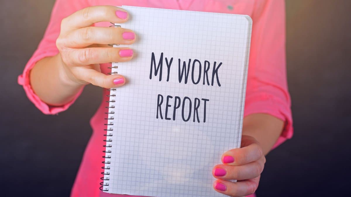 A woman in a pink shirt holds a notebook labeled 'My Work Report' indoors, showcasing text.