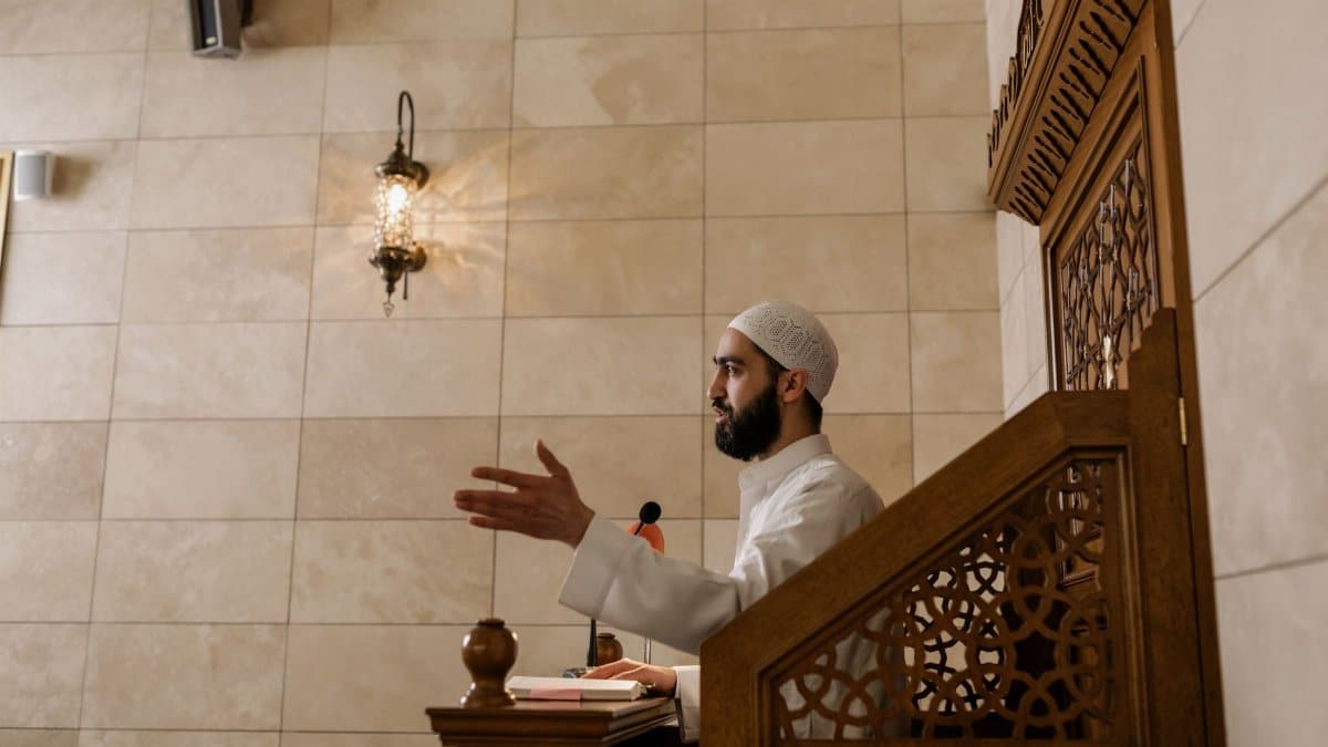 An imam delivering a sermon from a wooden lectern inside a beautifully decorated mosque.