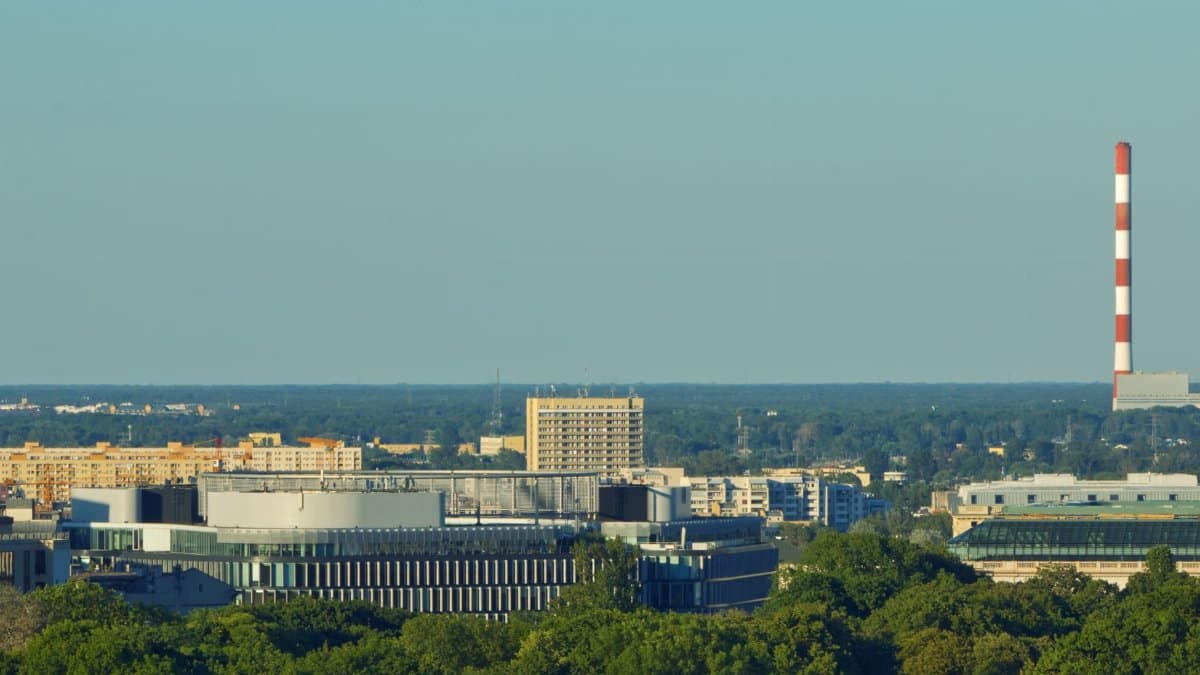 A wide-angle view of a city skyline featuring industrial buildings and a striped smokestack against a clear blue sky.