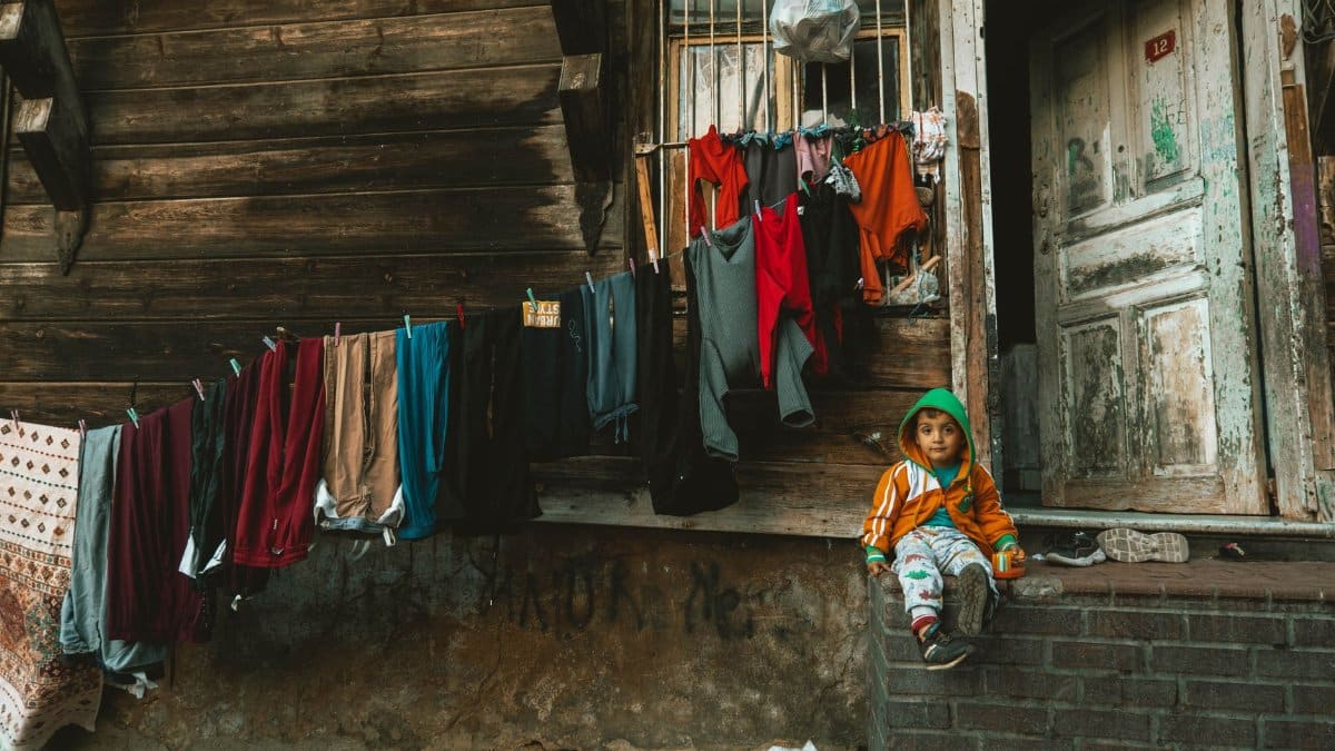 A child in a colorful hoodie sits outside a rustic wooden house in Istanbul. Clothes hang to dry.