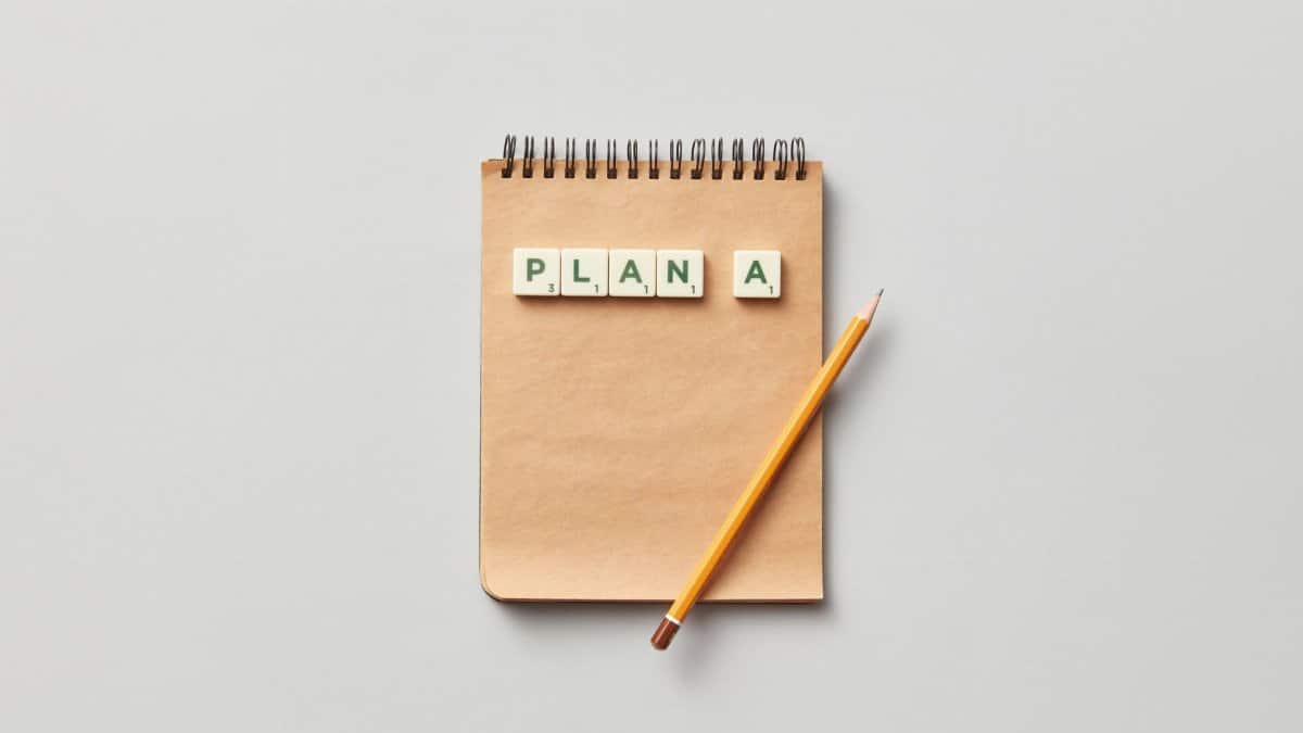 Top view of a notepad with a pencil and scrabble tiles spelling 'PLAN A' on a gray background.
