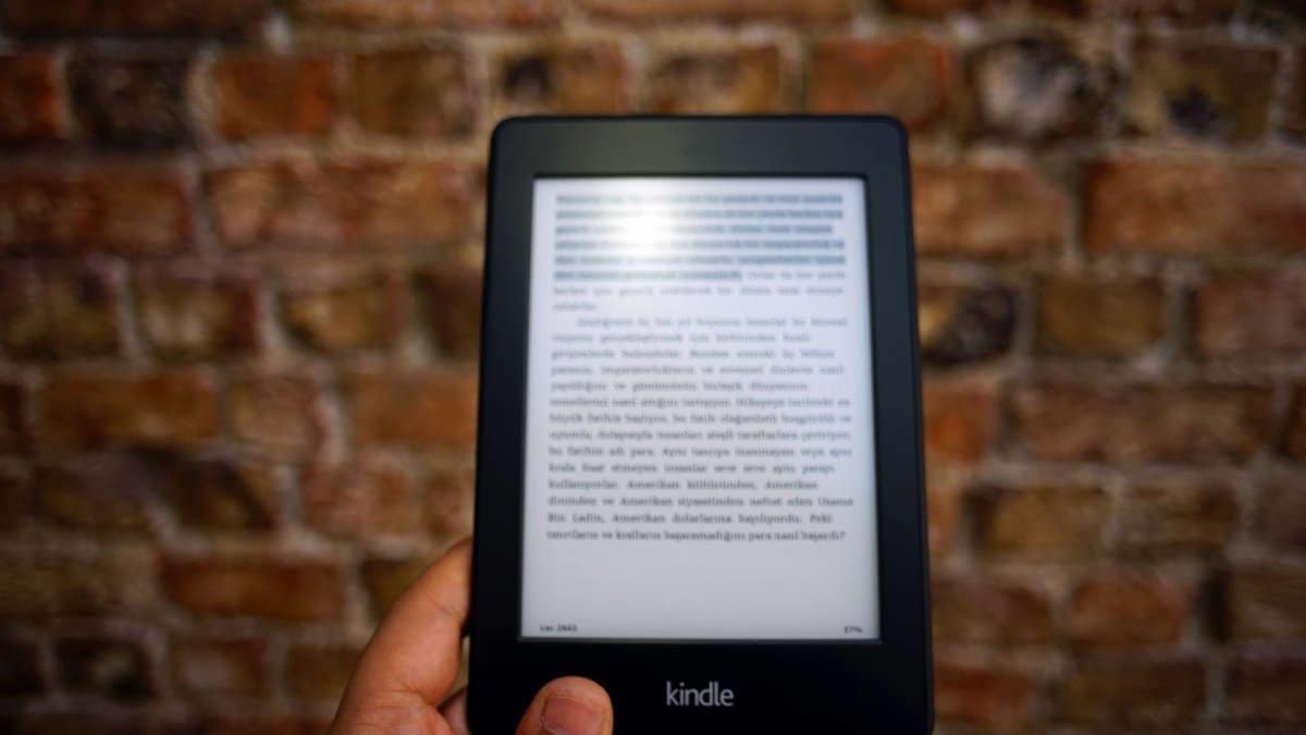 Close-up of a Kindle e-reader held by a person, showcasing a digital reading device against a textured brick wall backdrop.