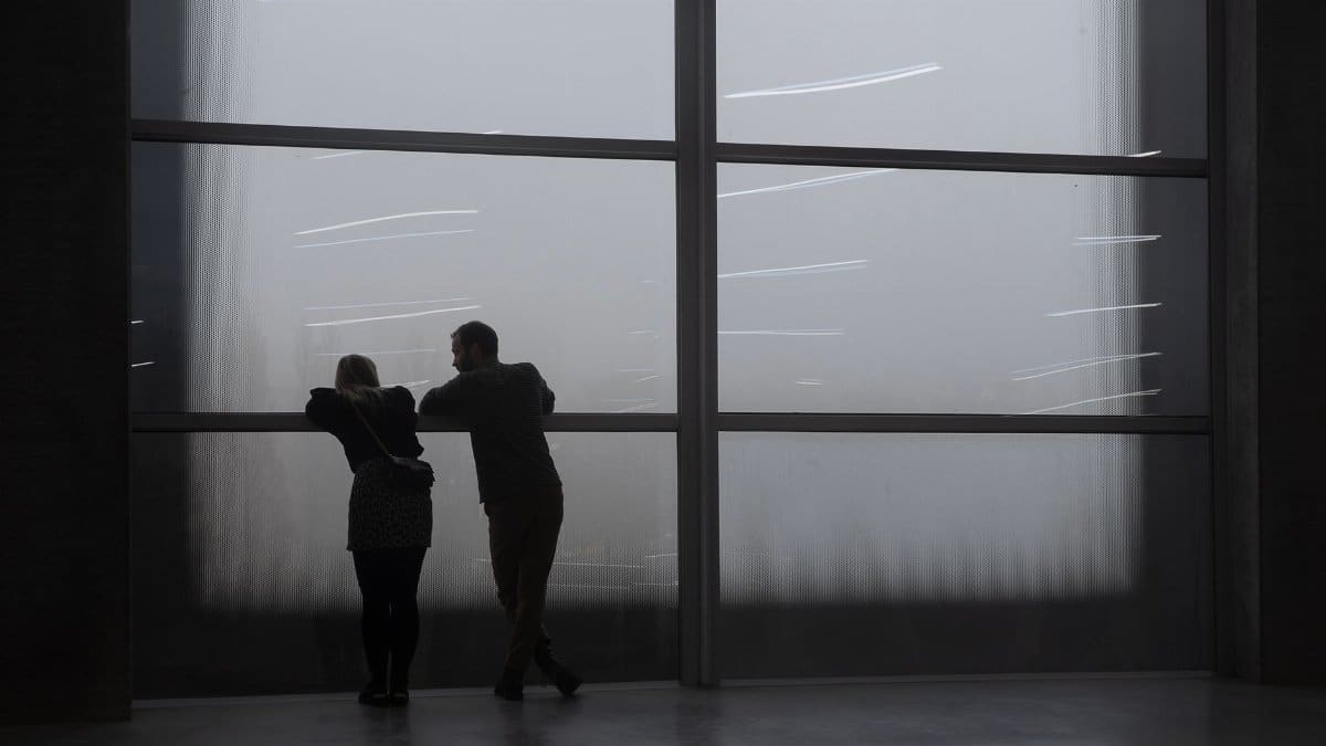 Silhouetted couple observes the foggy cityscape through a large window indoors.
