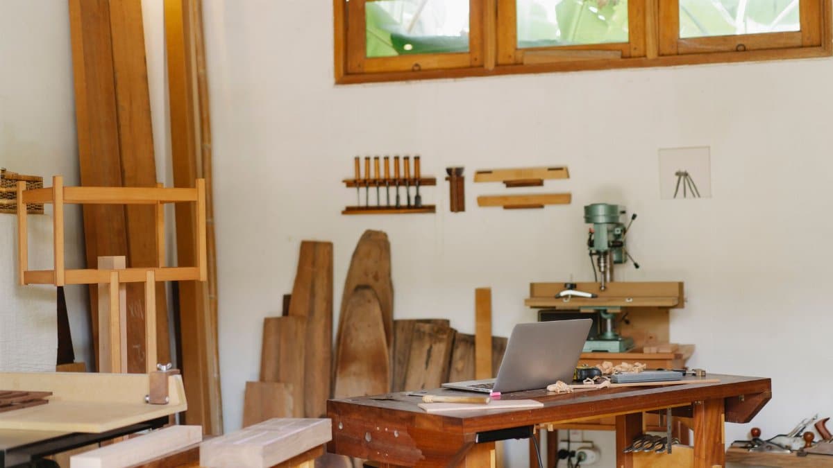 Opened laptop placed on table in traditional carpentry workshop full of wooden boards and tools in sunlight