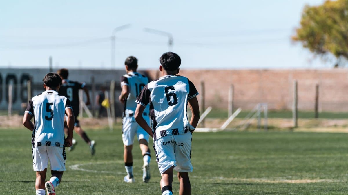Teen boys playing soccer on a green field under a clear blue sky, wearing team jerseys.