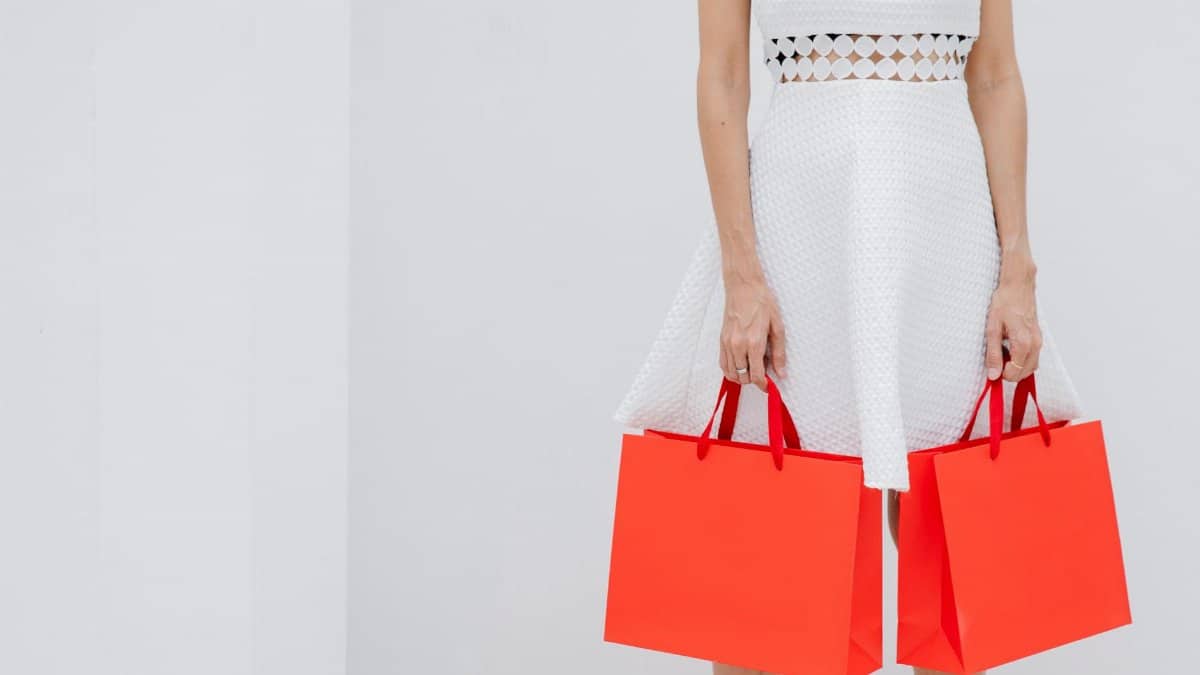 Crop anonymous female in stylish white dress carrying red paper shopping bags against light background in studio