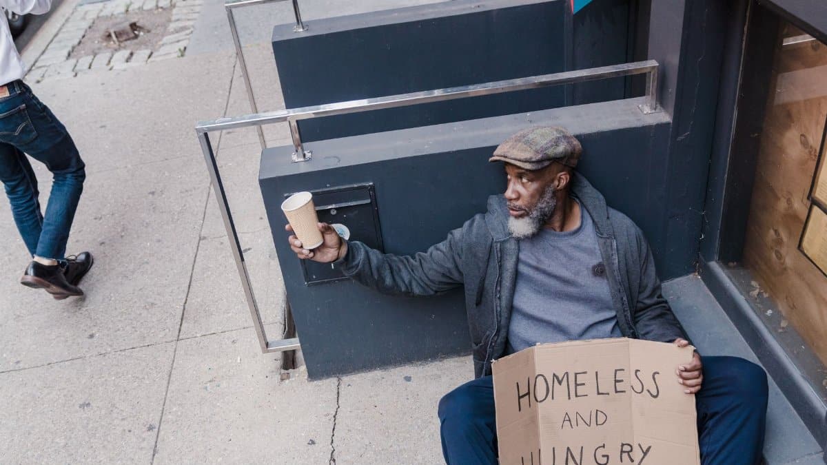 A homeless man sitting on a sidewalk holding a cup and sign in an urban area.