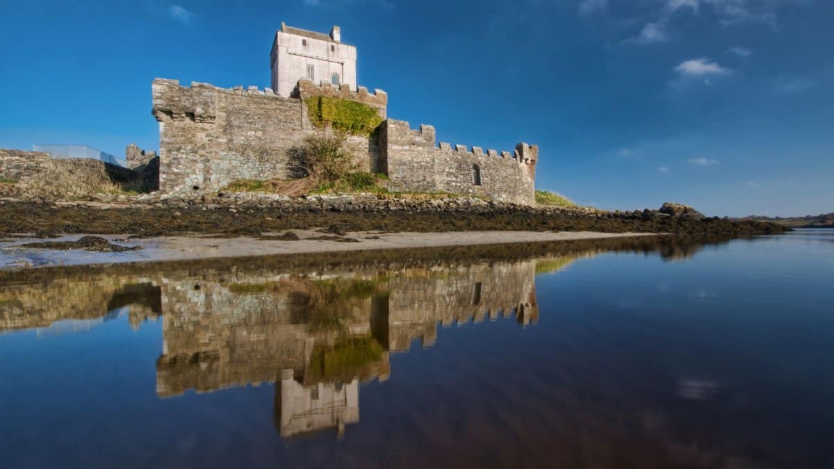 A stunning view of Doe Castle in County Donegal, Ireland reflecting in calm waters under a clear blue sky.