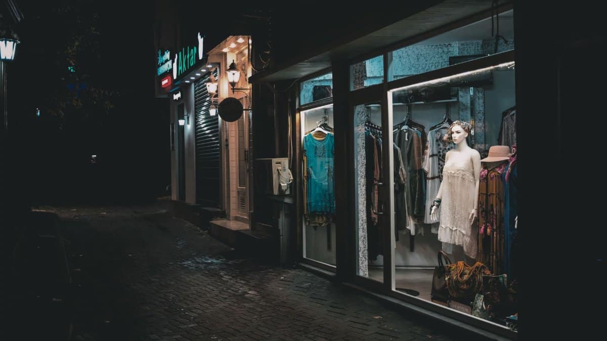 A boutique's fashion display brightly lit in a dark alley at night, showcasing elegant dresses.