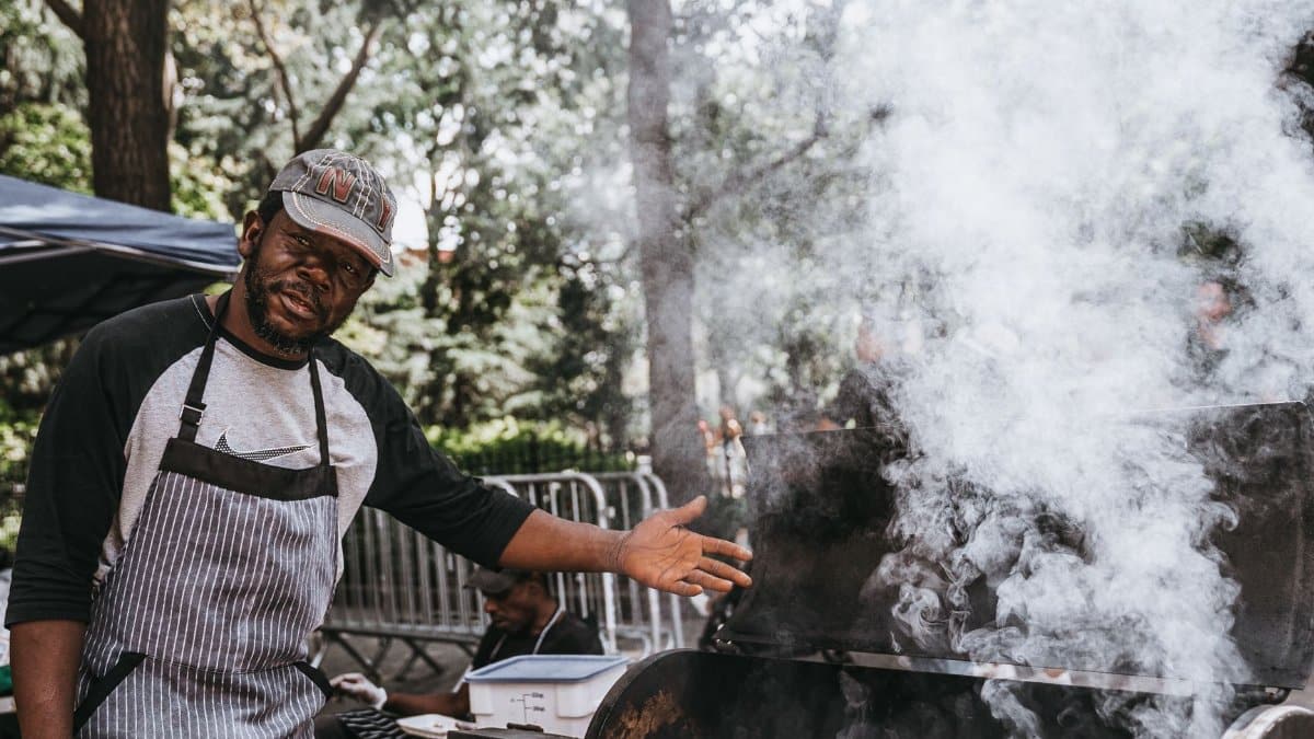 A man grilling meat outdoors with smoke rising from the barbecue.