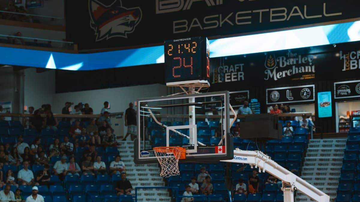 Close-up of a basketball hoop inside a stadium with a scoreboard and audience in the background.