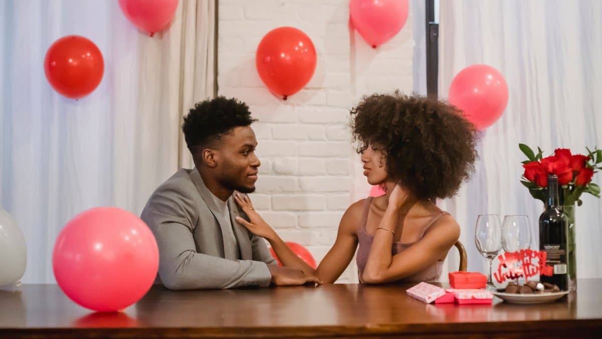Young African American couple in elegant outfits in room at table with wineglasses and bottle with drink near bouquet of roses in vase with gifts and looking at each other