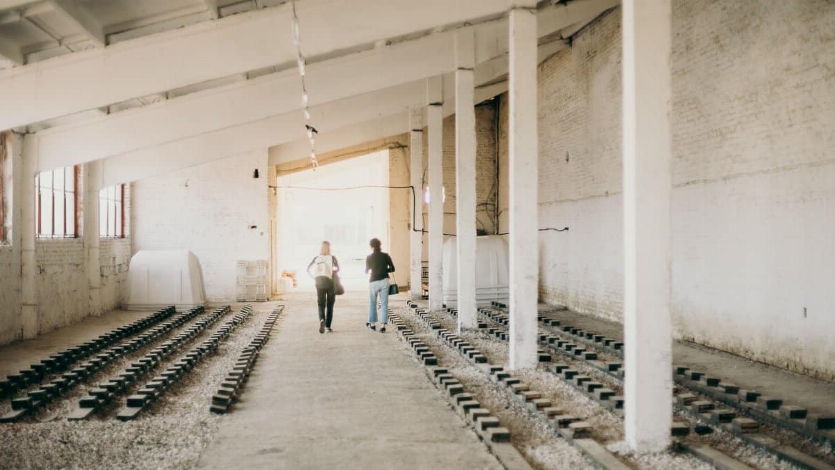 Two people walking inside a spacious warehouse with industrial rail tracks and structural columns.