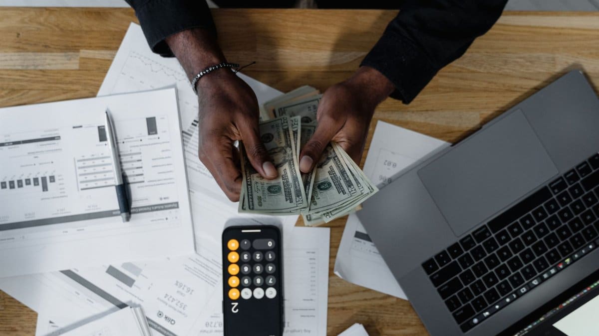 Person counting US dollars, using a calculator and laptop, with financial documents on a wooden desk.