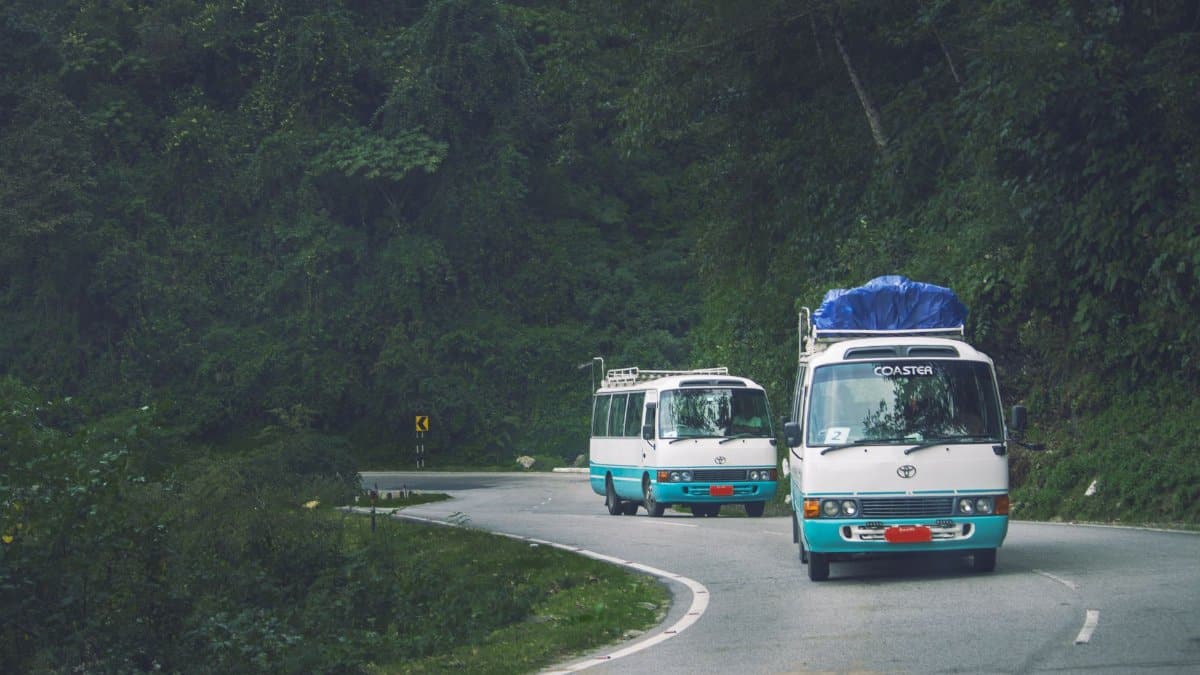Two mini buses travel a scenic mountain road in Phuentsholing, Bhutan, surrounded by lush green forests.