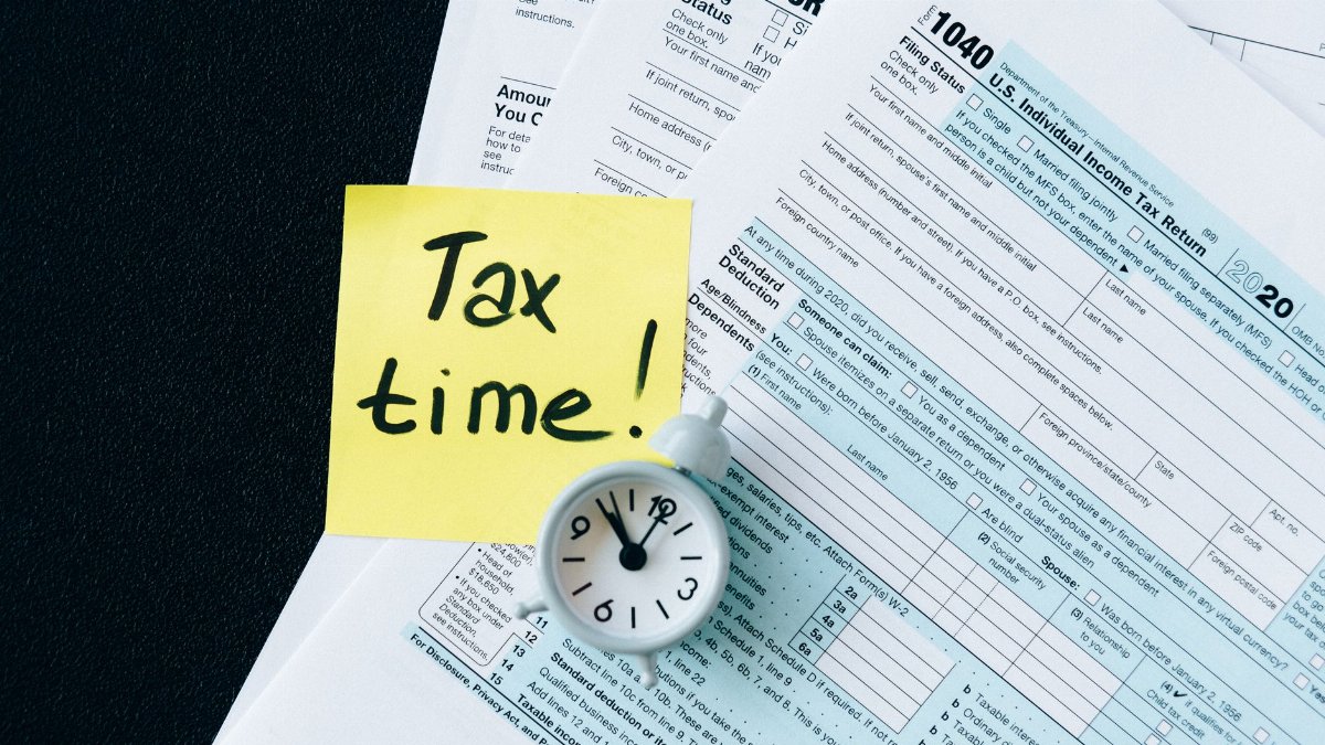 A stack of tax forms with a clock and yellow sticky note saying 'Tax time!' indicating urgency.