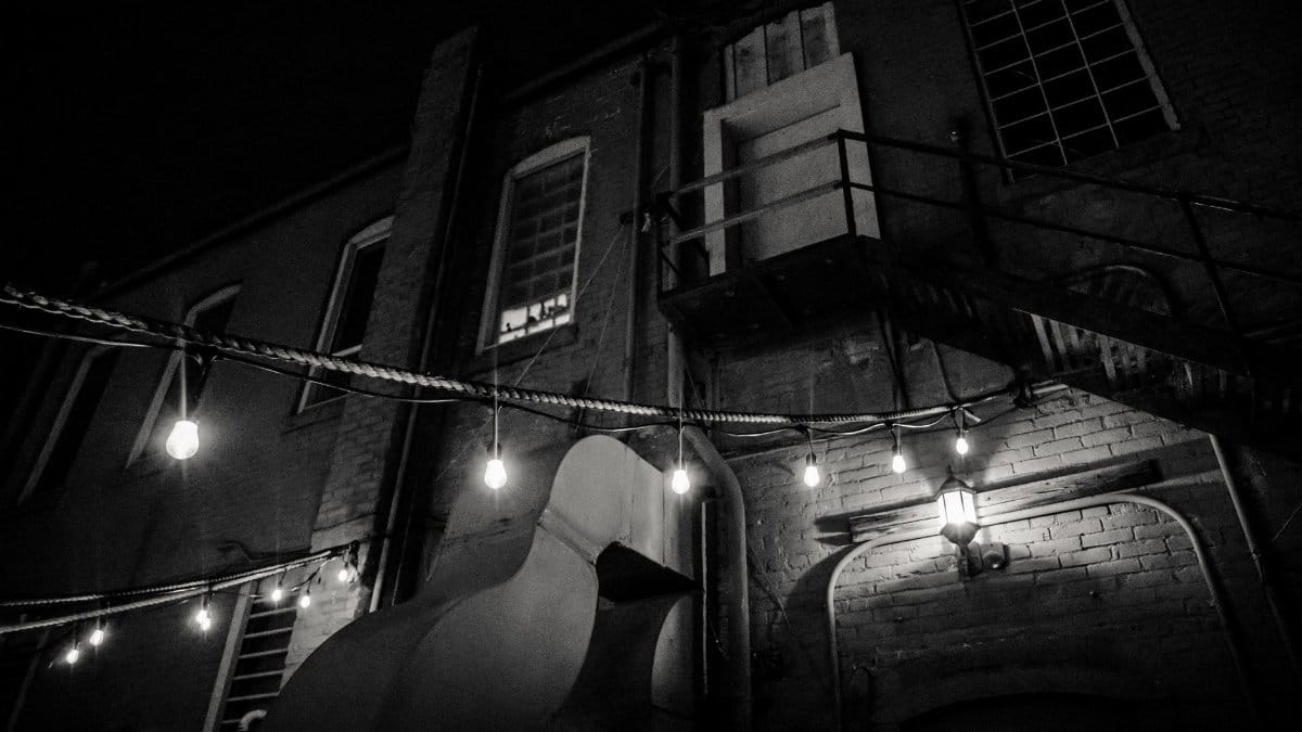 A dark and moody urban alleyway illuminated by string lights, captured in grayscale.