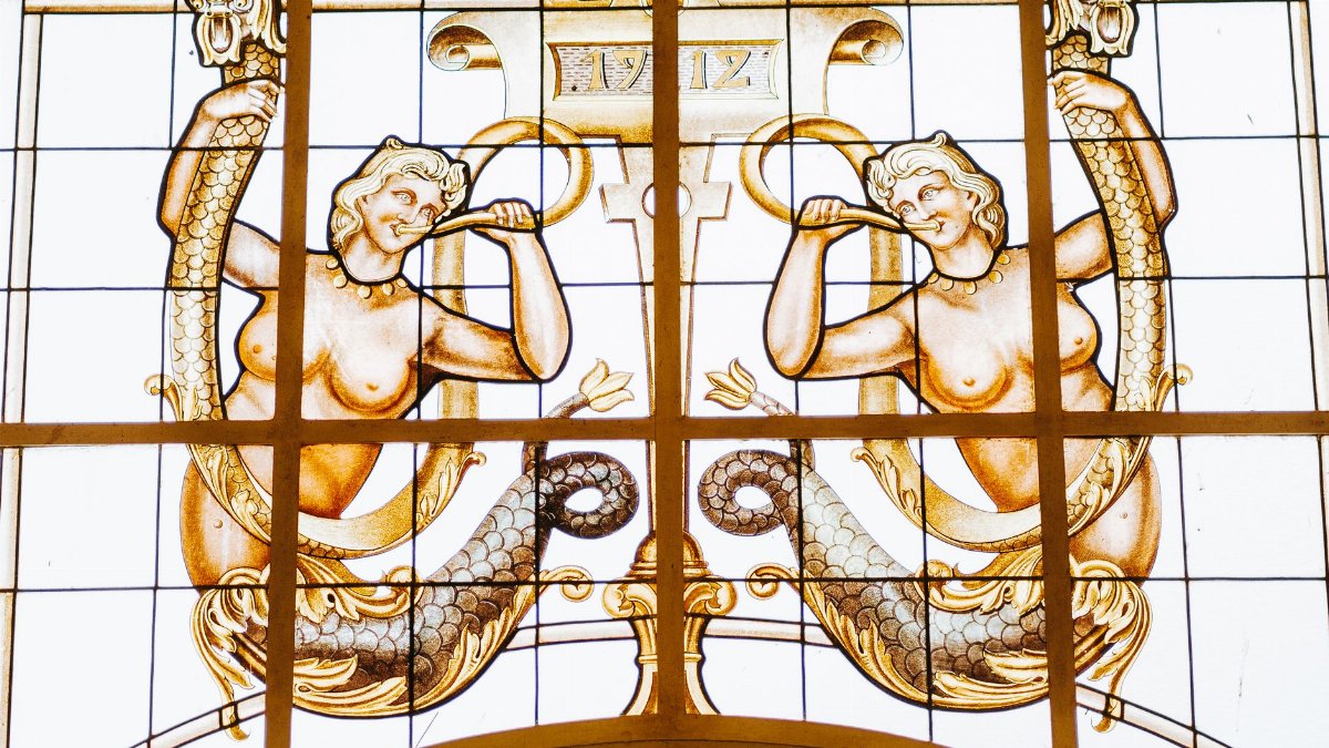 Ornate stained glass window depicting mythical sirens playing trumpets in Caxambu, Brazil.