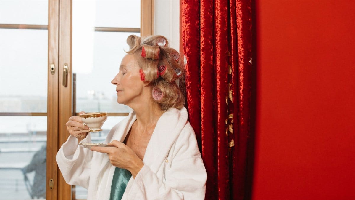 Relaxed senior woman in bathrobe with curlers, savoring tea by the window.