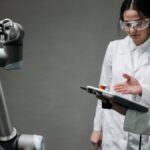 Researchers in lab coats and safety glasses engaging with a robotic arm in a lab setting.