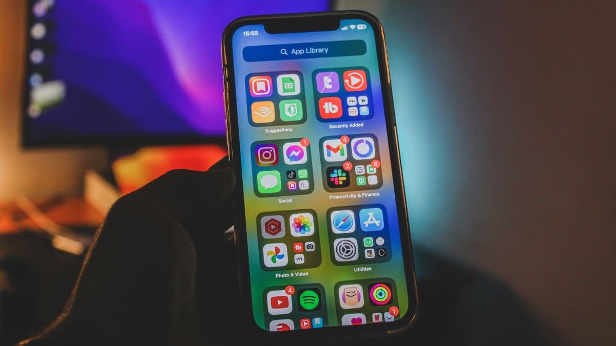 A hand holds a smartphone showing a colorful app-filled screen in an indoor room with dim lighting.