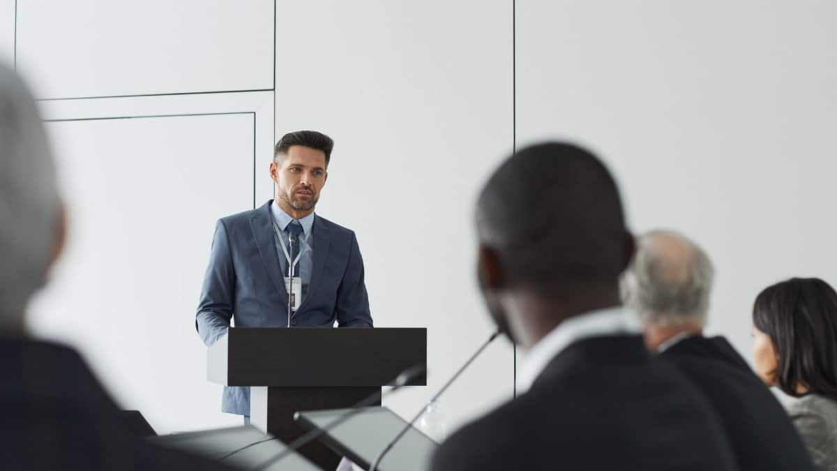 Speaker at a conference delivering a presentation to a diverse audience in a modern setting.