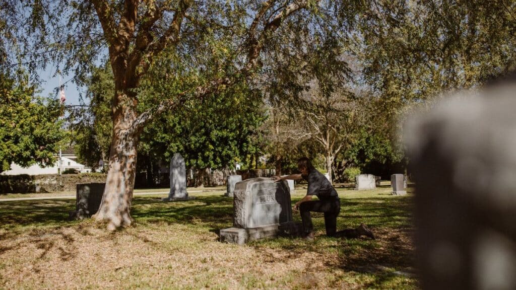 A man kneels by a grave in a sunlit cemetery, reflecting moments of grief and solitude.