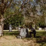 A man kneels by a grave in a sunlit cemetery, reflecting moments of grief and solitude.