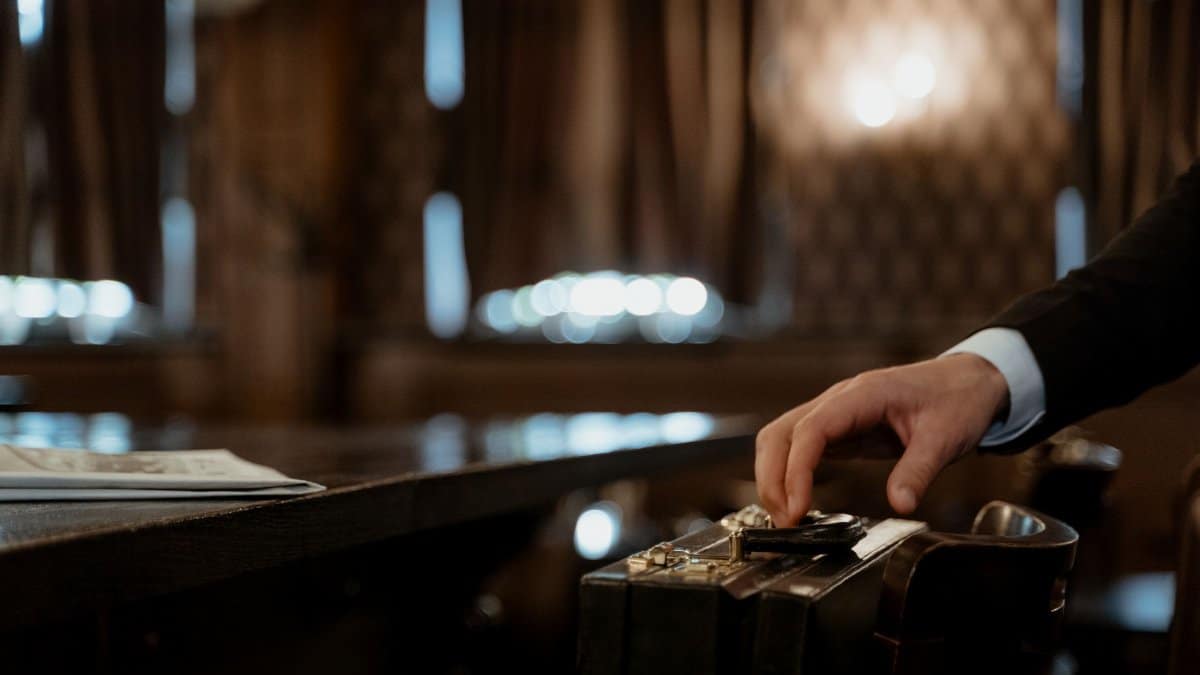 A close-up shot captures a hand holding a black briefcase in a dimly lit elegant room with a blurred background.